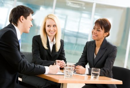 three people wearing suits sitting around a small table and talking to each other