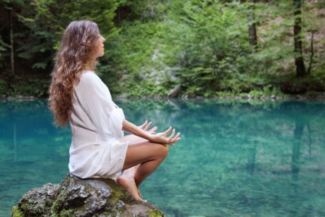 woman wearing white sitting on a rock meditating by a river