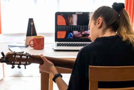 a woman plays an acoustic guitar while studying music theory guitar on her laptop