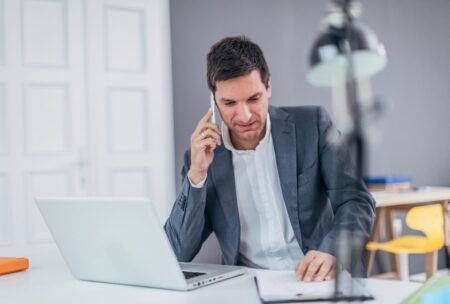 A man in a suit sits at a desk, using a laptop and talking on the phone. He is engaged in cold calling.