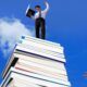 A man stands triumphantly on a pile of books, symbolizing the wisdom and knowledge needed to succeed in business.