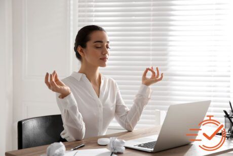 A woman sits at her desk, meditating with her laptop, embracing mindfulness in her work environment.