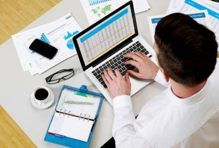 a man focused on his laptop, surrounded by papers and items, working on his personal financial statement