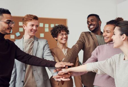 a diverse group of individuals stands united, hands together in front of a board, symbolizing pronouns and intersectionality